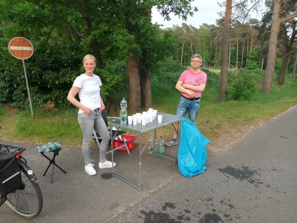 Zwei Personen stehen an einem Tisch neben einer Straße im Wald, der mit Getränken und Bechern gedeckt ist. Im Hintergrund sind Bäume und ein Verkehrsschild zu sehen.