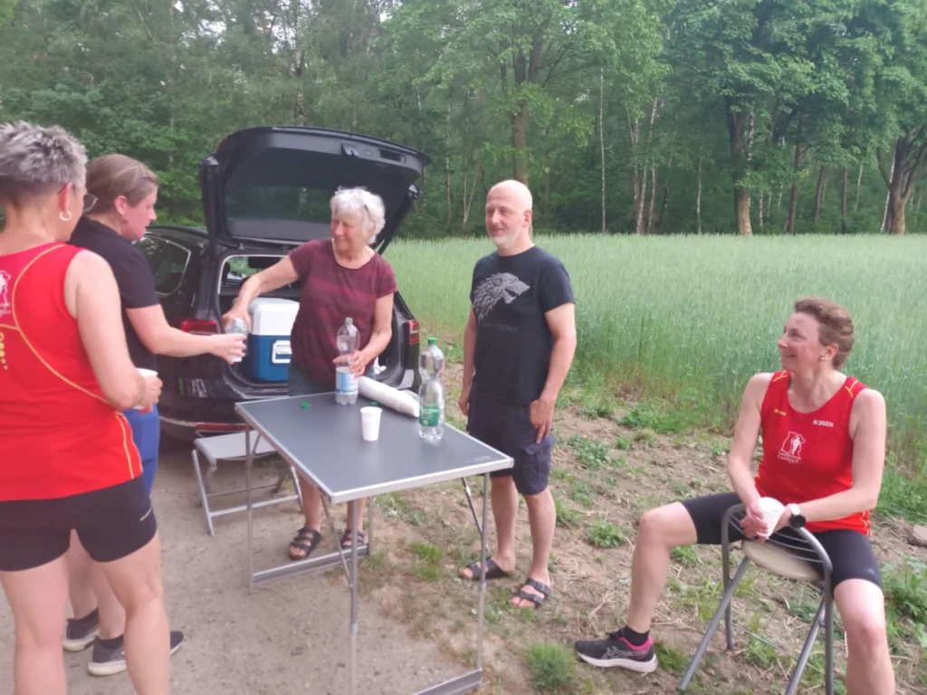 Gruppe von fünf Personen bei einer Outdoor-Veranstaltung, die Getränke aus einem Auto und einem Tisch servieren. Umgeben von einer grünen Wiese und Bäumen, entspannen sie nach einer sportlichen Aktivität.