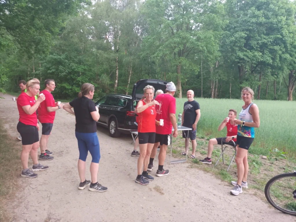 Gruppe von Läufern in roten T-Shirts, die nach einem Training im Wald stehen und Getränke genießen. Im Hintergrund steht ein Auto und es sind grüne Bäume und Wiesen zu sehen.