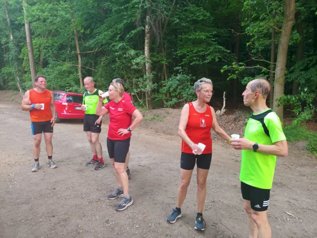 Gruppe von Läufern in Sportkleidung, die sich im Wald unterhalten und Getränke halten, während ein rotes Auto im Hintergrund steht. Geeignet für Themen wie Freizeitsport, Laufen und Gruppentraining.