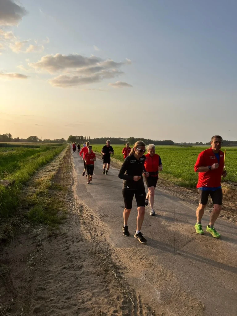 Gruppe von Läufern in roten Shirts, die auf einem ländlichen Weg bei Sonnenuntergang joggen, umgeben von grünen Feldern und blauen Himmel.
