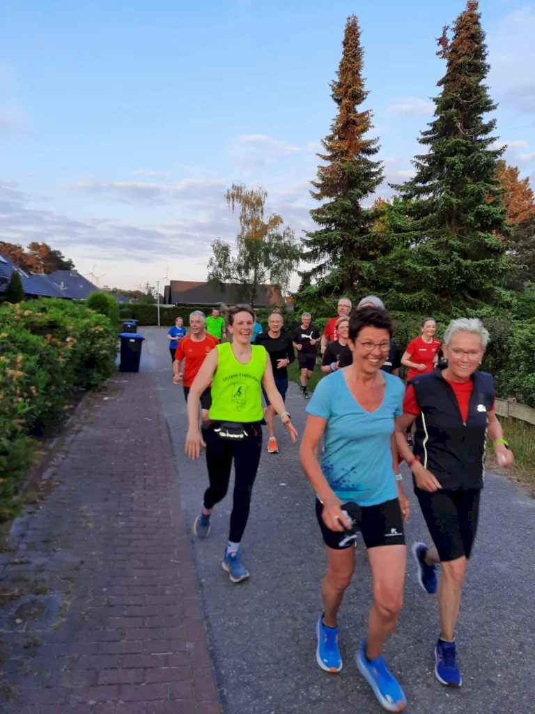 Gruppe von Läufern in sportlicher Kleidung, die auf einem Weg joggen, umgeben von Bäumen und Wohnhäusern. Die Atmosphäre ist lebhaft und freundlich, ideal für gemeinsames Laufen und Fitness.