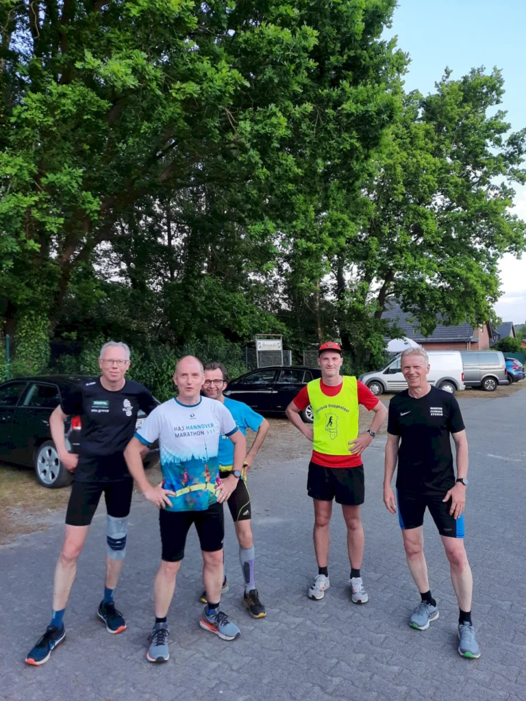 Fünf Männer in Sportbekleidung stehen auf einem Parkplatz unter Bäumen, bereit für ein Lauftraining. Im Hintergrund sind Autos und ein Schild sichtbar.