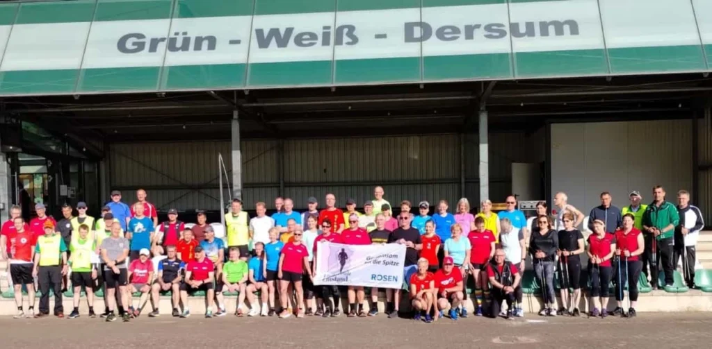 Gruppenfoto von Läufern und Sportbegeisterten vor dem Gebäude des Vereins Grün-Weiß-Dersum, mit verschiedenen Trikots in bunten Farben und einem Banner, das die Gemeinschaft und den Zusammenhalt symbolisiert.