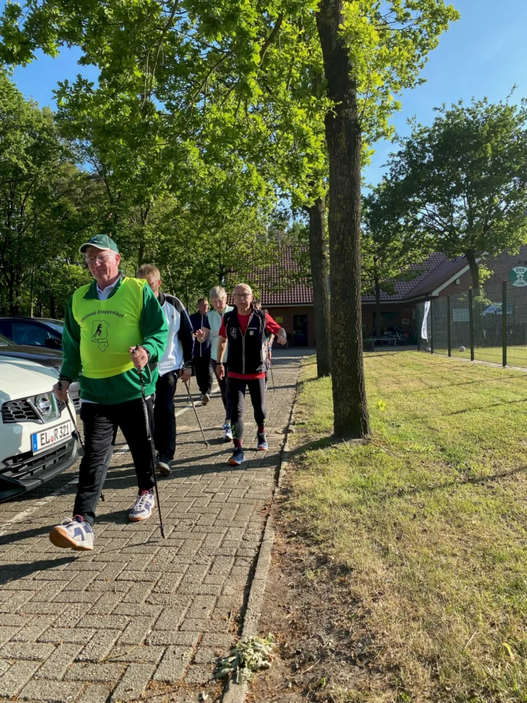 Gruppe von Senioren beim Nordic Walking auf einem gepflasterten Weg, umgeben von Bäumen und Grünflächen, mit einem Gebäude im Hintergrund.
