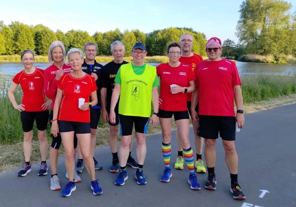 Gruppe von Läufern in sportlicher Kleidung am Ufer eines Gewässers, lächelnd und posierend für das Foto, während sie an einer Laufveranstaltung teilnehmen. Hintergründe zeigen grüne Bäume und einen klaren Himmel.