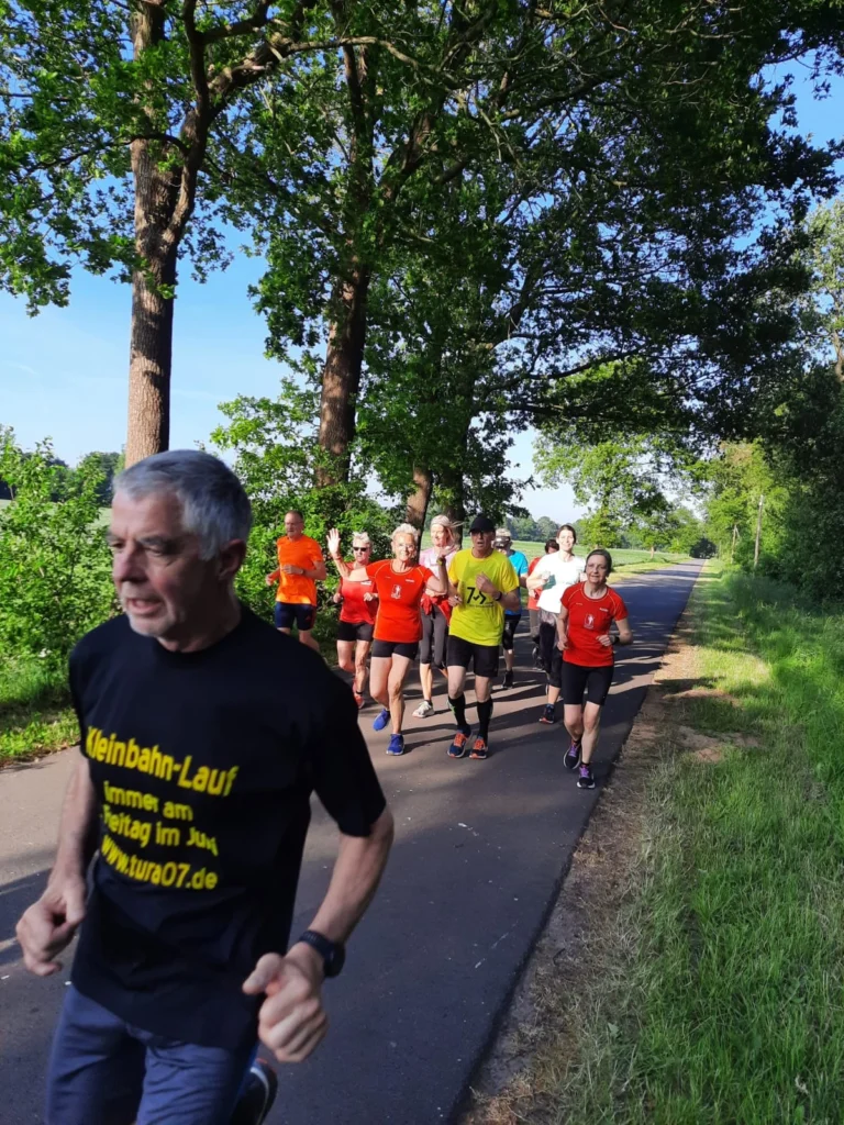 Gruppentraining beim Kleinbahn-Lauf, Teilnehmer in bunten Laufshirts joggen auf einem schattigen Weg umgeben von Bäumen, sonniger Tag im Juni.