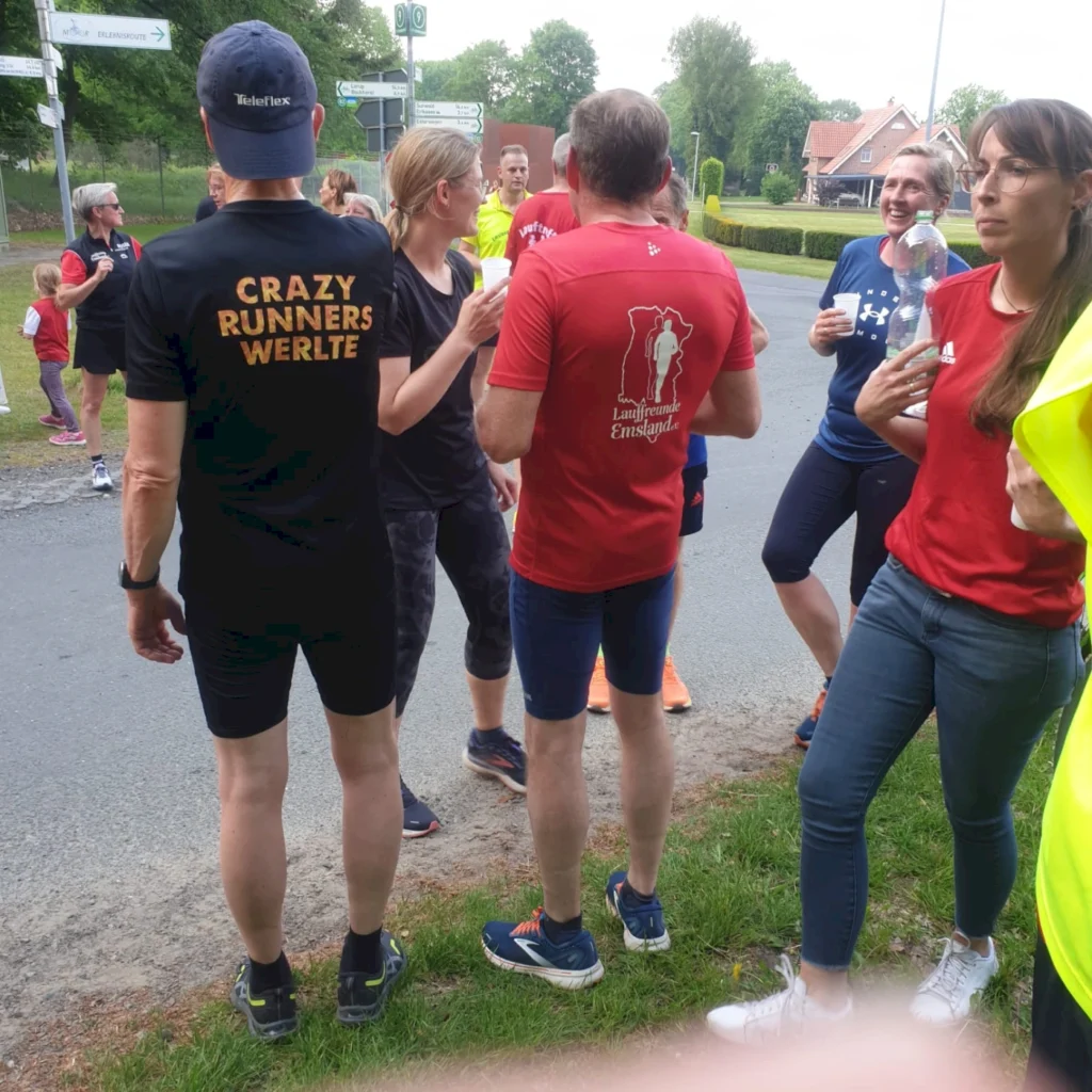 Gruppe von Läufern bei einem Event in Werlte, einige tragen T-Shirts mit Aufdrucken wie "Crazy Runners" und "Laufende Freunde Emsland", während sie sich unterhalten und Wasser trinken.