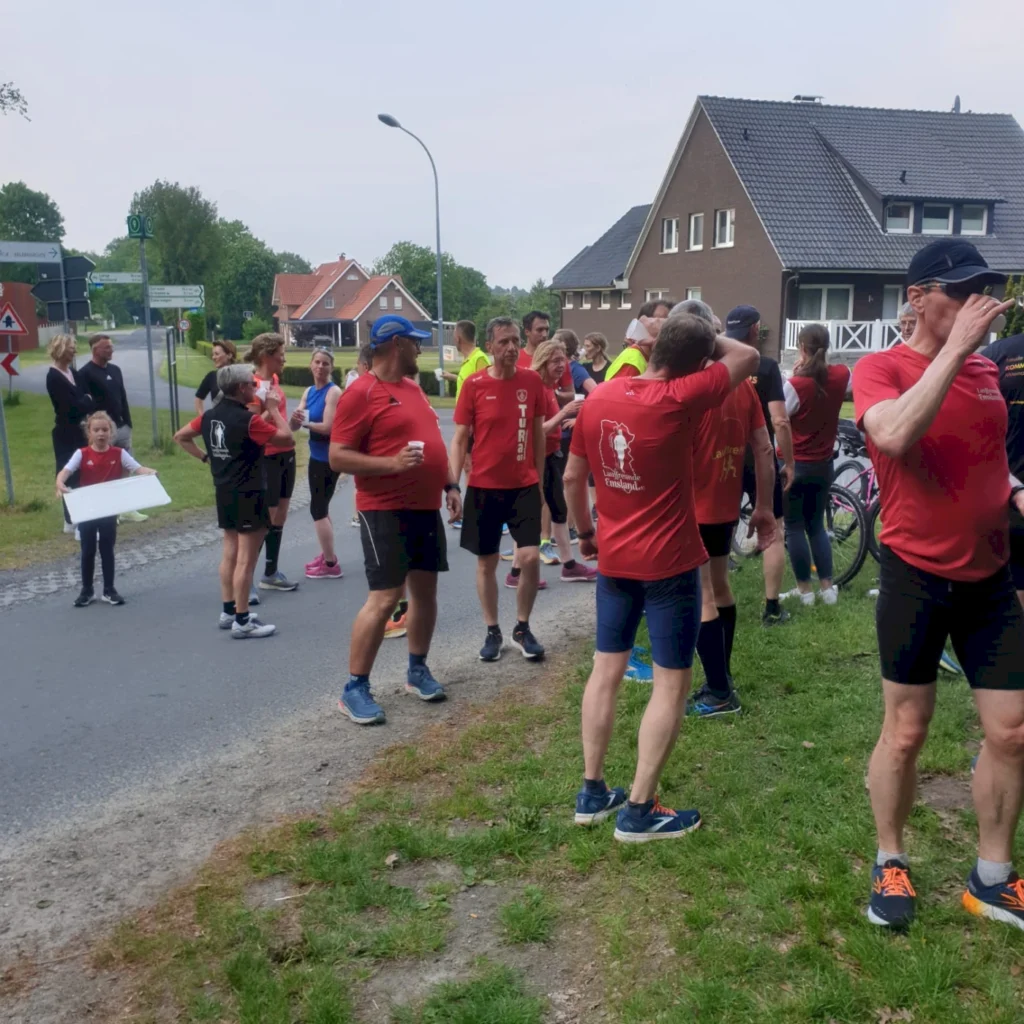 Gruppierung von Läufern in roten Shirts während einer Laufveranstaltung, mit Zuschauern und einer Person, die ein Schild hält, an einer Straße in einer ländlichen Umgebung.
