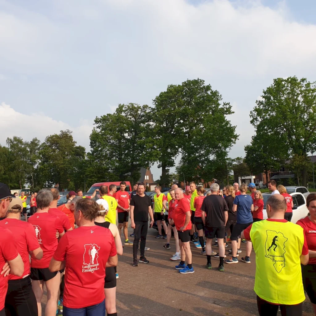 Gruppierung von Läufern in roten und gelben T-Shirts bei einem Lauf-Event in Emsland, umgeben von Bäumen und Autos, an einem sonnigen Tag.