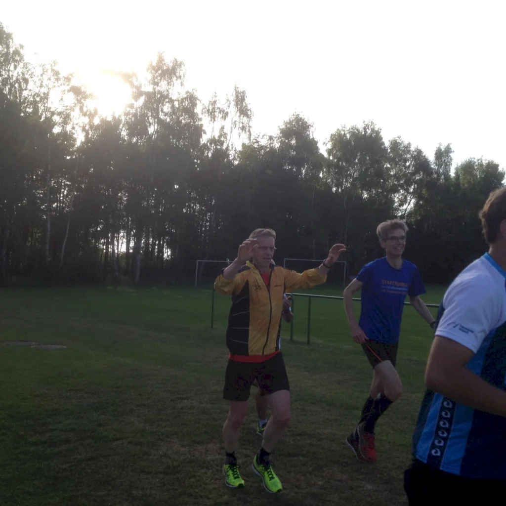 Zwei Läufer in Sportkleidung joggen auf einem Fußballplatz, umgeben von Bäumen. Der Mann in der gelben Jacke hebt seine Hände, während der andere Läufer in einem blauen Shirt entspannt läuft. Die Sonne scheint im Hintergrund.