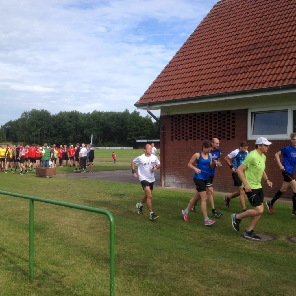 Läufer in Sportbekleidung joggen auf einem Stadiongelände, während im Hintergrund eine Gruppe von Zuschauern steht. Der Himmel ist bewölkt, und das Gelände ist gut gepflegt.
