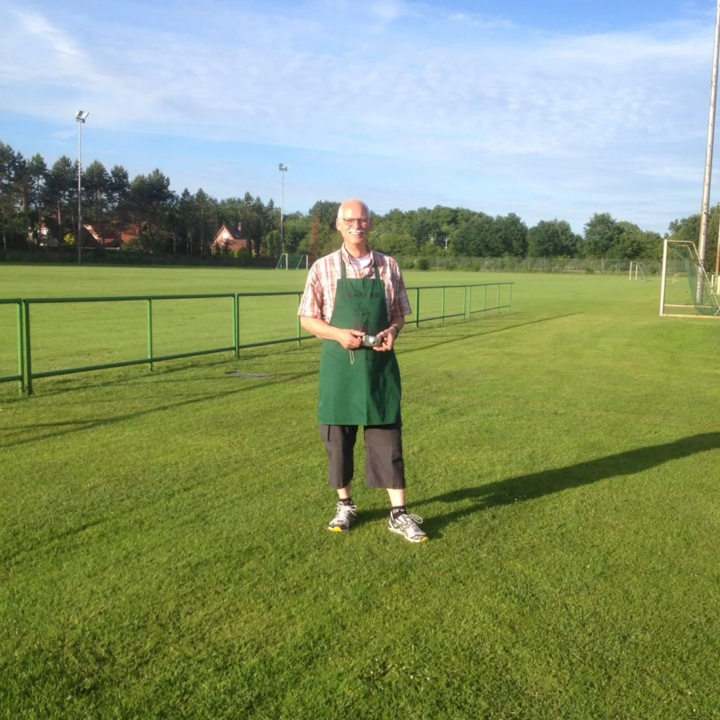 Mann mit Brille und grünem Schürze steht auf einem gepflegten Fußballplatz, umgeben von grüner Wiese und Bäumen, unter blauem Himmel mit wenigen Wolken.