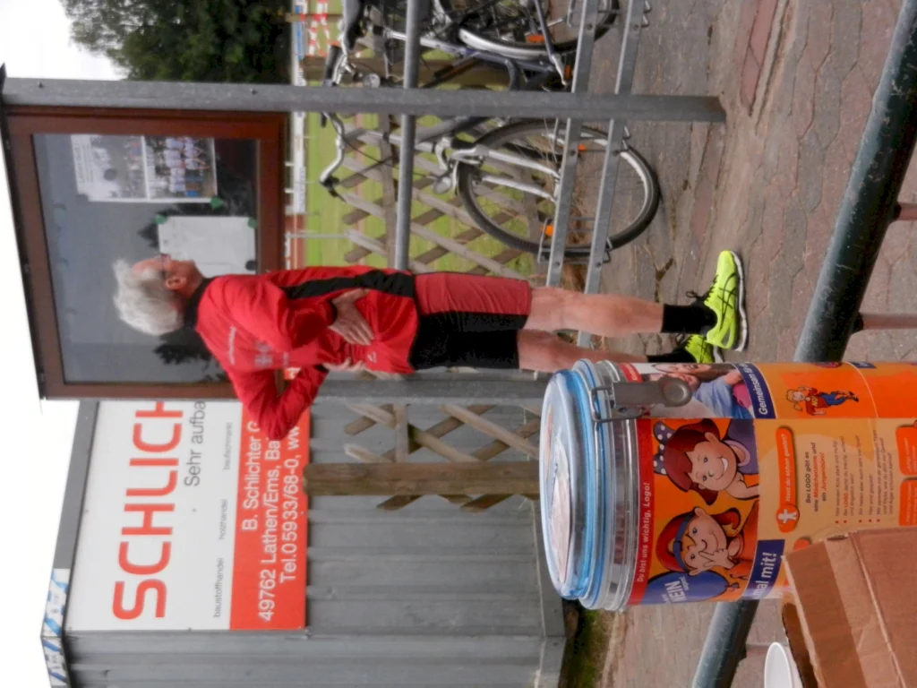 Ein älterer Mann in roter Sportbekleidung steht vor einem Informationsschild. Im Hintergrund sind Fahrräder abgestellt, und ein farbenfroher Behälter mit kindgerechten Motiven ist im Vordergrund sichtbar.