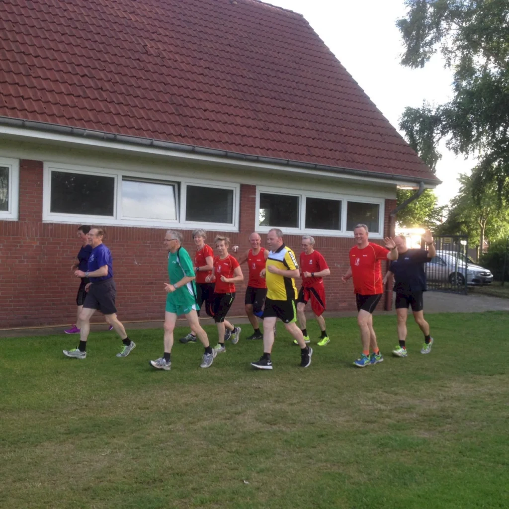 Eine Gruppe von zehn älteren Menschen joggt in Sportkleidung auf einem Rasen neben einem Gebäude mit Fenstern. Die Teilnehmer tragen verschiedene Farben, darunter Rot, Grün und Schwarz, und scheinen aktiv und motiviert zu sein.