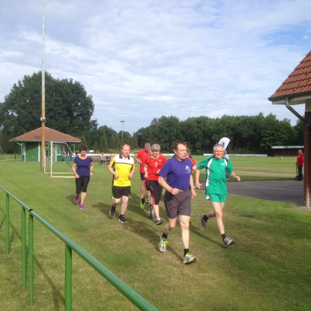 Gruppe von Läufern unterschiedlichen Alters und Geschlechts, die auf einem Sportplatz joggen, umgeben von grünem Gras und Bäumen, bei klarem Himmel.