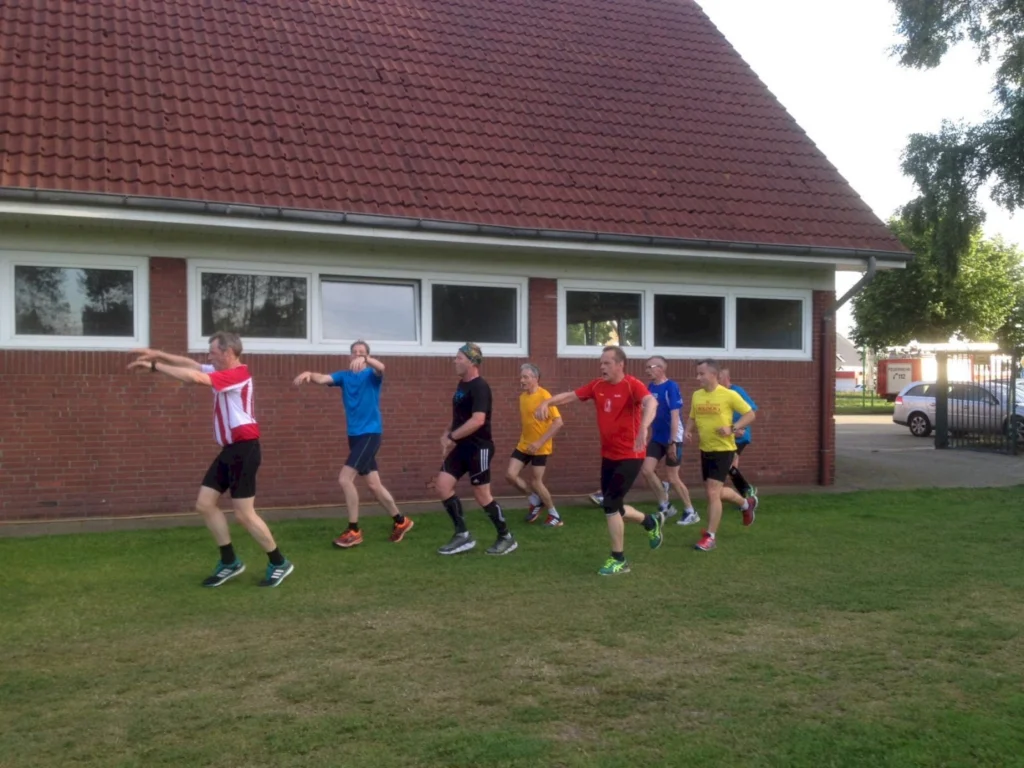 Gruppe von sieben Männern in Sportkleidung, die im Freien auf einer Wiese vor einem Gebäude joggen. Sie tragen unterschiedliche T-Shirts in leuchtenden Farben und zeigen verschiedene Laufstile.