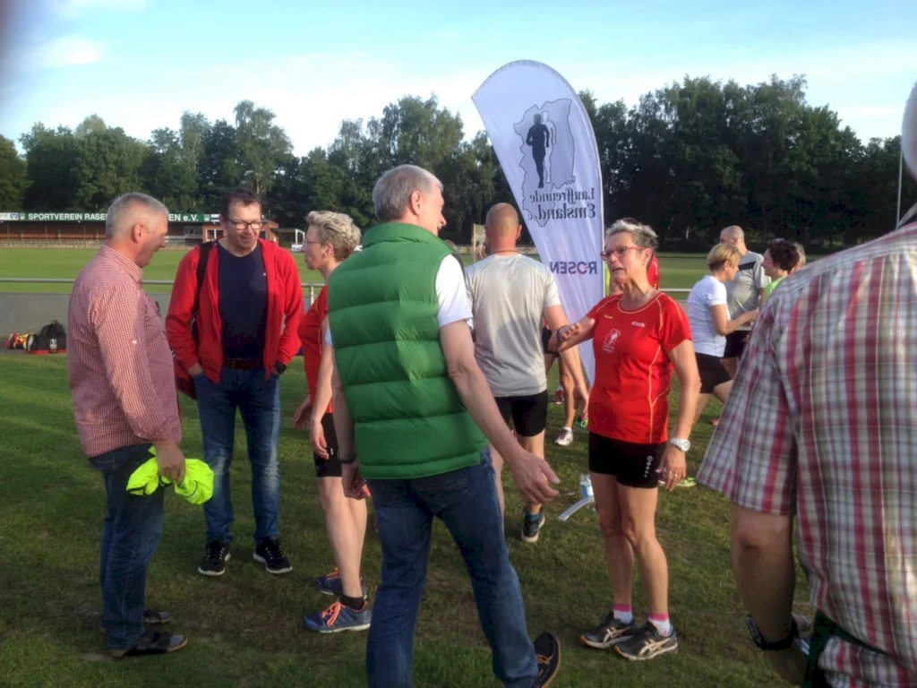 Gruppe von Läufern und Unterstützern beim Sportverein auf einem Rasenplatz, umgeben von Bäumen. Im Vordergrund stehen zwei Männer und eine Frau im Gespräch, während im Hintergrund weitere Teilnehmer aktiv sind. Ein Werbebanner für den Laufveranstalter ist sichtbar.