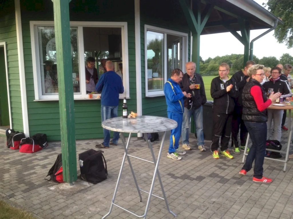 Menschen stehen an einem Imbissstand in einem Sportverein, während sie Snacks und Getränke genießen. Im Vordergrund ist ein Stehtisch zu sehen, im Hintergrund wird Essen ausgegeben. Die Umgebung ist grün und einladend, ideal für gesellige Zusammenkünfte nach sportlichen Aktivitäten.