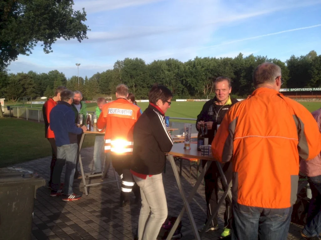 Menschen stehen an Stehtischen auf einem Sportplatz, einige tragen Feuerwehrjacken. Im Hintergrund sind Fußballfelder und Bäume zu sehen. Es scheint eine gesellige Veranstaltung zu sein, bei der Getränke serviert werden.