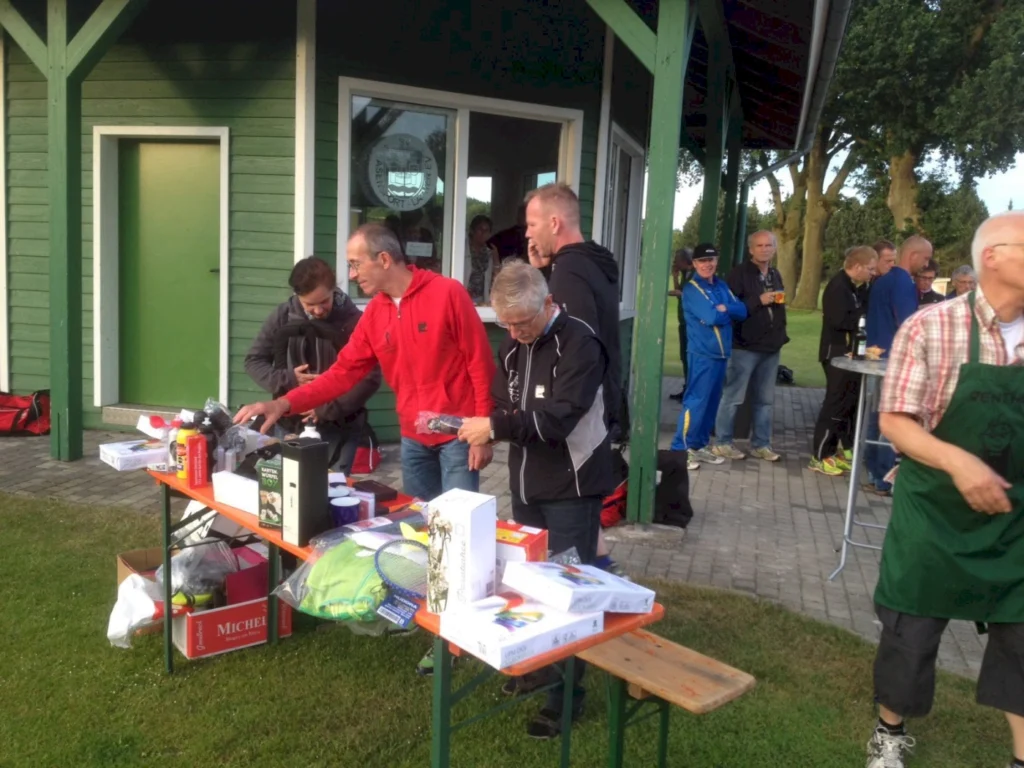 Menschen stehen an einem Tisch mit Sportartikeln und Geschenken im Freien, während sie an einem Vereinsgebäude in einer Sportanlage teilnehmen. Im Hintergrund sind weitere Personen zu sehen, die sich unterhalten und die Veranstaltung genießen.