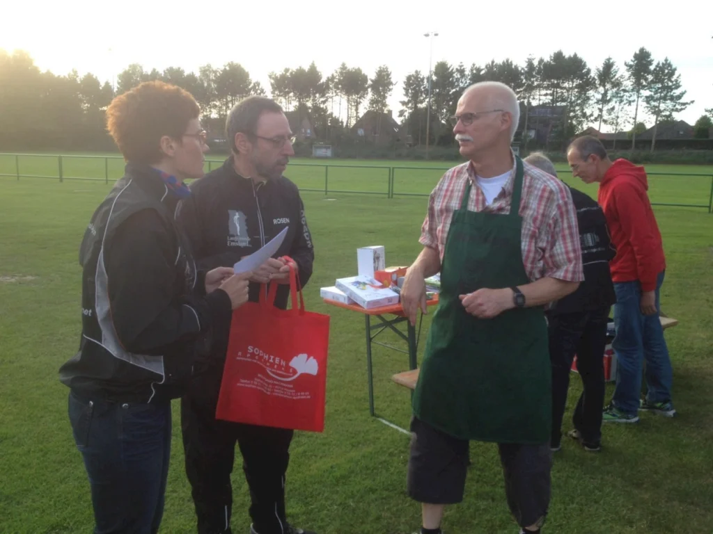 Personen bei einer Veranstaltung auf einem Sportplatz, die sich um einen Tisch mit verschiedenen Artikeln versammeln. Eine Frau mit lockigem Haar spricht mit einem Mann, während ein weiterer Mann in Schürze und Brille im Hintergrund steht. Die Atmosphäre ist freundlich und gemeinschaftlich.