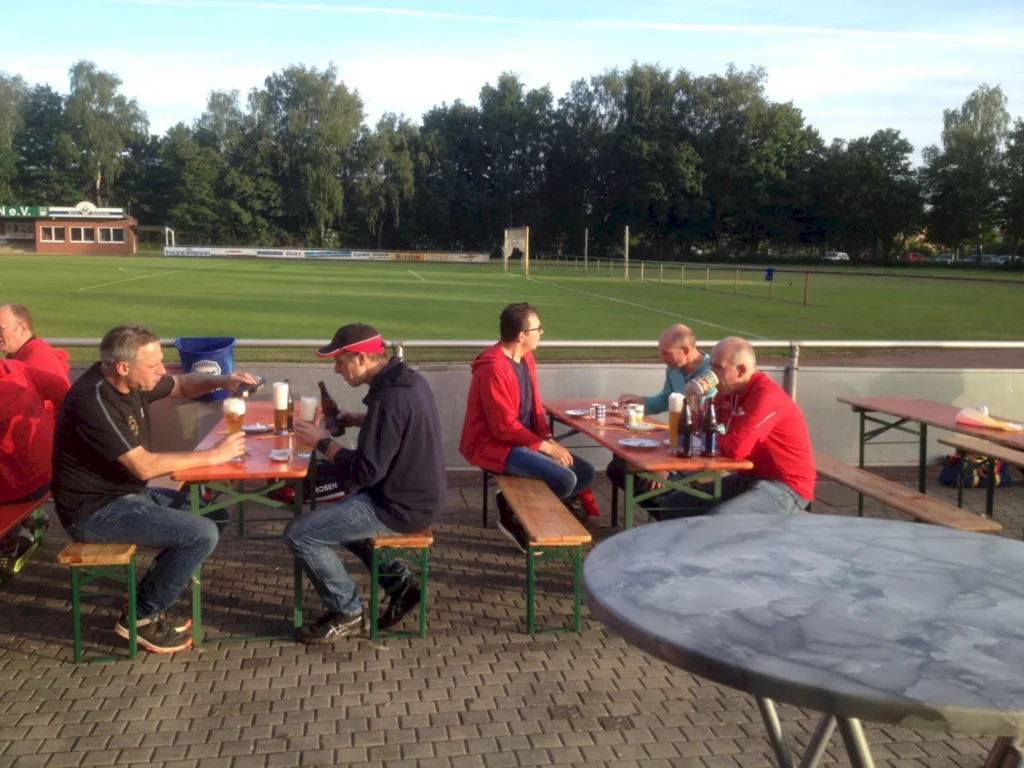Menschen sitzen an Tischen im Freien und genießen Getränke, während sie auf einem Sportplatz entspannen. Die Szenerie zeigt eine gesellige Atmosphäre mit einem Fußballfeld im Hintergrund.