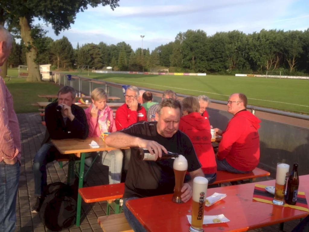Menschen sitzen an Tischen in einem Freiluftbereich, genießen Bier und Snacks, während sie ein Fußballspiel im Hintergrund beobachten. Die Atmosphäre ist entspannt und gesellig, mit einer grünen Sportplatzlandschaft und Bäumen im Hintergrund.