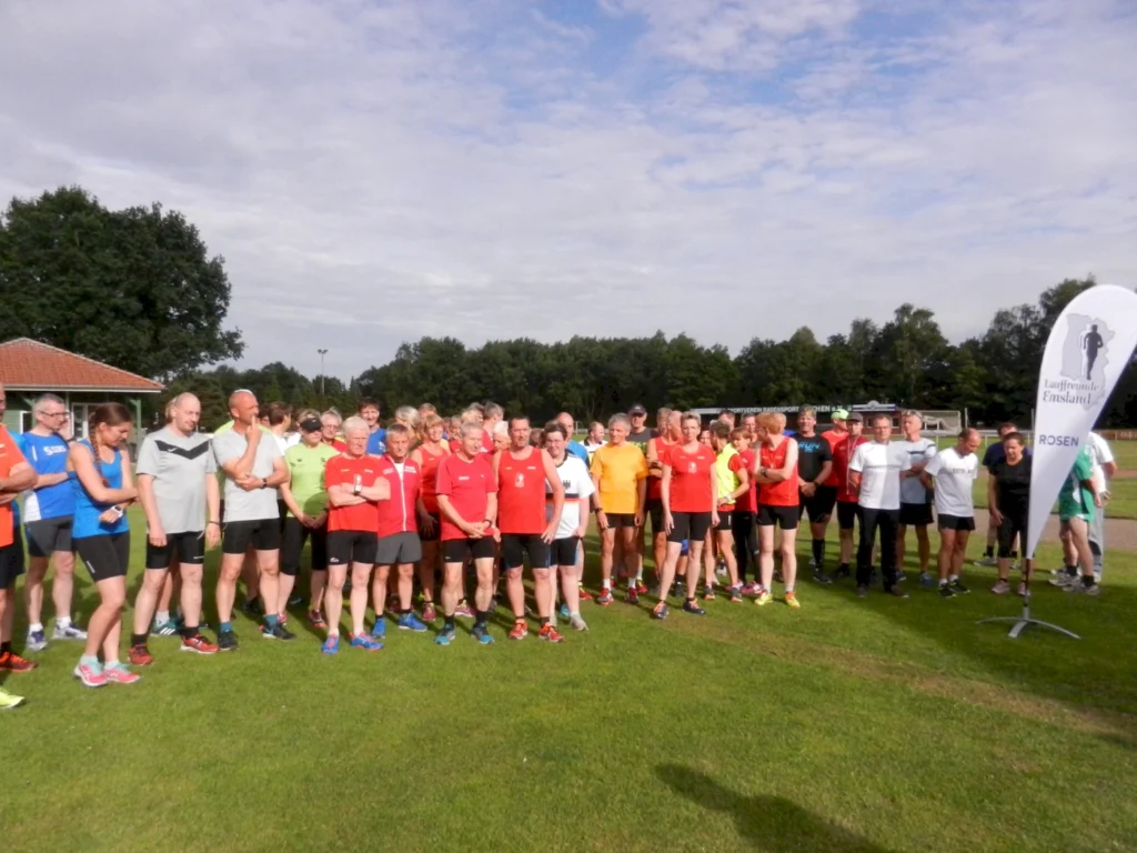 Gruppenfoto von Laufbegeisterten beim Sportevent, auf einem Sportplatz in Deutschland. Teilnehmer in verschiedenen Laufoutfits stehen auf einer Wiese, umgeben von Bäumen und einem Vereinsbanner im Hintergrund.