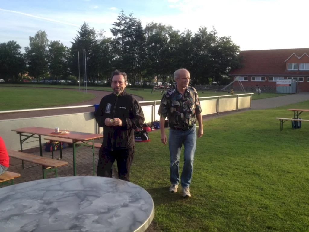 Zwei Männer stehen auf einem Sportplatz, umgeben von Bäumen und einem Gebäude im Hintergrund. Der Mann links trägt eine schwarze Jacke mit dem Schriftzug "ROSEN" und hält etwas in der Hand, während der Mann rechts ein buntes, bedrucktes Hemd trägt und auf den Betrachter zugeht. Im Vordergrund ist ein runder Tisch aus Marmor zu sehen.