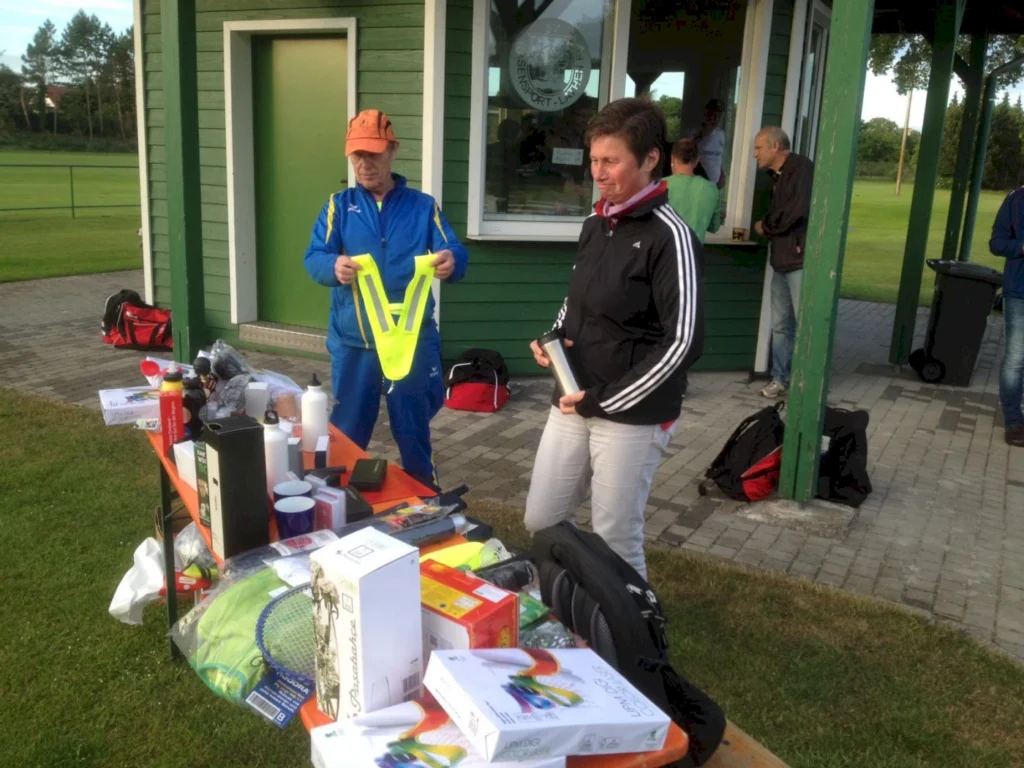 Zwei Personen stehen an einem Tisch mit Sportartikeln und Zubehör, während im Hintergrund ein Vereinsgebäude zu sehen ist. Der Mann in blauer Sportkleidung hält eine gelbe Warnweste, während die Frau in schwarzer Jacke eine Flasche in der Hand hält. Der Tisch ist mit verschiedenen Gegenständen wie Schlägern, Getränken und weiteren Sportutensilien bedeckt.