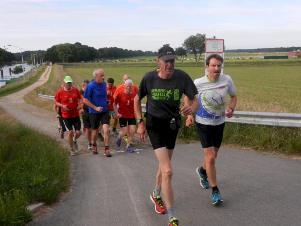 Gruppe von Läufern in Sportkleidung, die einen Weg entlanglaufen, umgeben von Feldern und Bäumen. Die Teilnehmer scheinen an einem Trainingslauf teilzunehmen, wobei einige rote T-Shirts tragen. Im Hintergrund ist ein Fluss sichtbar.