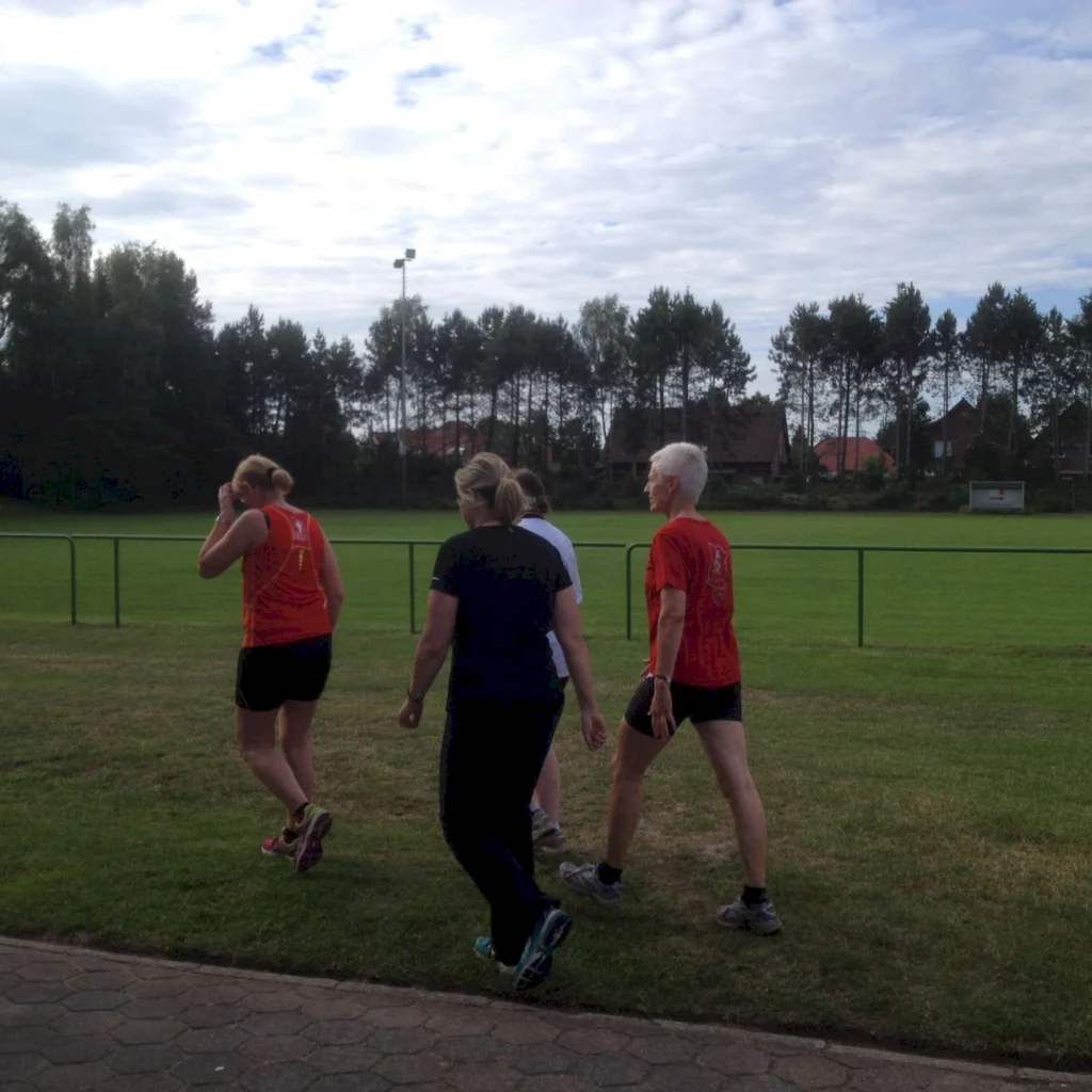 Vier Frauen in Sportkleidung gehen auf einem grünen Sportplatz, umgeben von Bäumen und einem klaren Himmel. Ideal für Fitness- und Laufaktivitäten.