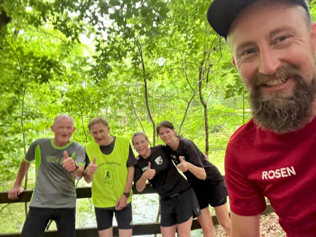 Gruppe von fünf Läufern, die im Wald posieren und Daumen hoch zeigen, während sie ihre Begeisterung für das Laufen und die Natur teilen.
