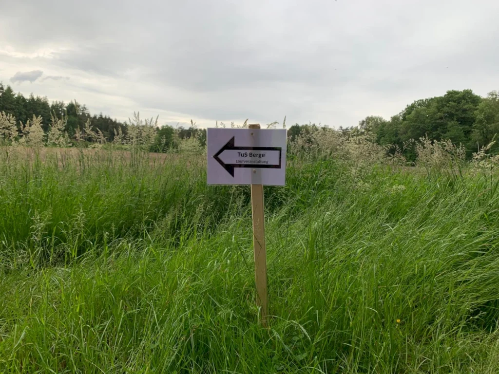 Schild mit Richtungspfeil nach links, das auf die Veranstaltung des TuS Berge hinweist, umgeben von hohem Gras und einer ländlichen Umgebung.