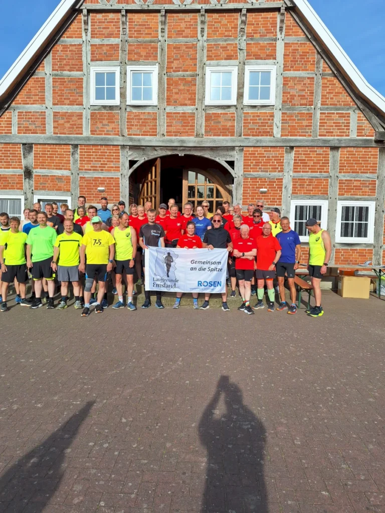 Gruppenfoto von Läufern in bunten Shirts vor einem historischen Fachwerkhaus, die ein Banner mit der Aufschrift "Gemeinsam an die Spitze" halten. Die Veranstaltung wird von den Laufenden Emsland organisiert.
