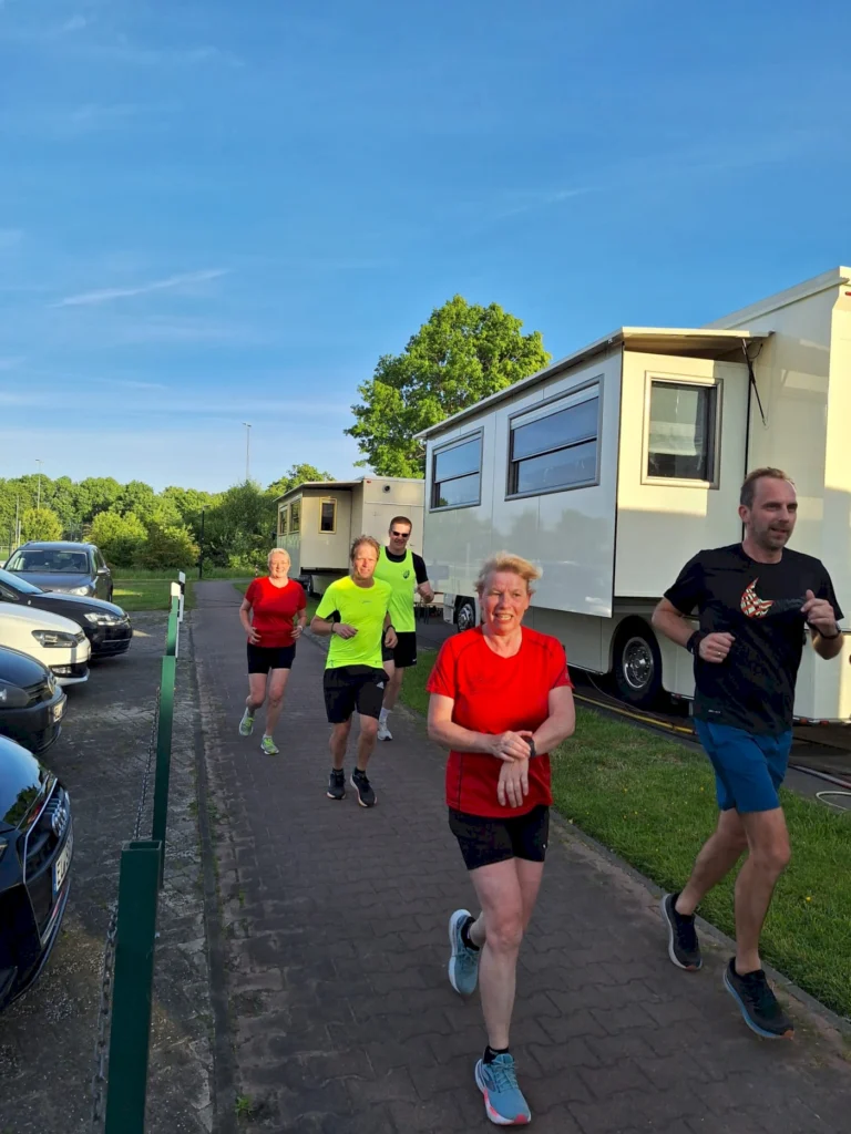 Gruppe von fünf Läufern in Sportkleidung, die auf einem Weg neben Wohnmobilen joggen. Die Szene zeigt eine sonnige Umgebung mit blauem Himmel und grüner Vegetation im Hintergrund.