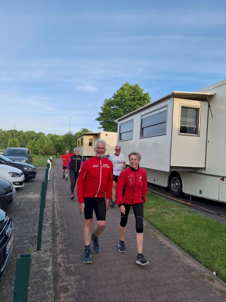 Gruppe von Menschen in Sportkleidung, die auf einem Gehweg neben Wohnmobilen spazieren. Im Hintergrund sind Bäume und ein blauer Himmel zu sehen. Ideal für Themen wie Outdoor-Aktivitäten und gesunde Lebensweise.