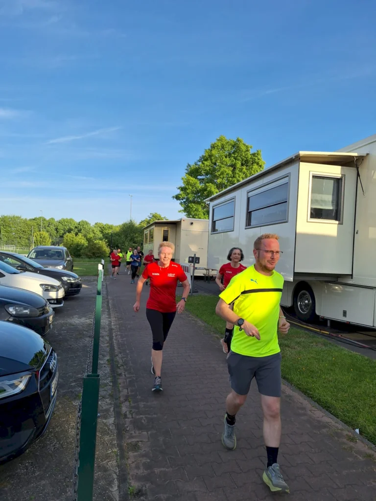 Gruppe von Läufern in Sportkleidung, die auf einem gepflasterten Weg entlang von Wohnwagen und parkenden Autos joggen, unter klarem Himmel und grüner Umgebung.