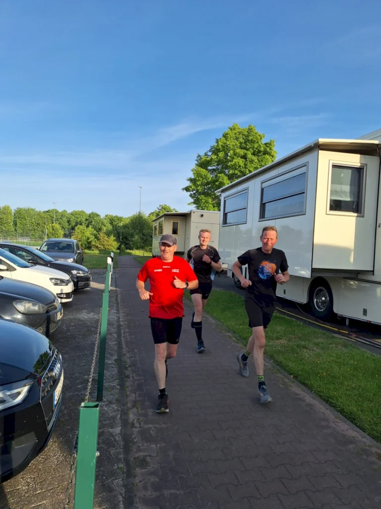 Drei Männer joggen auf einem Gehweg neben parkenden Autos und einem Wohnmobil, umgeben von grüner Landschaft und blauem Himmel.