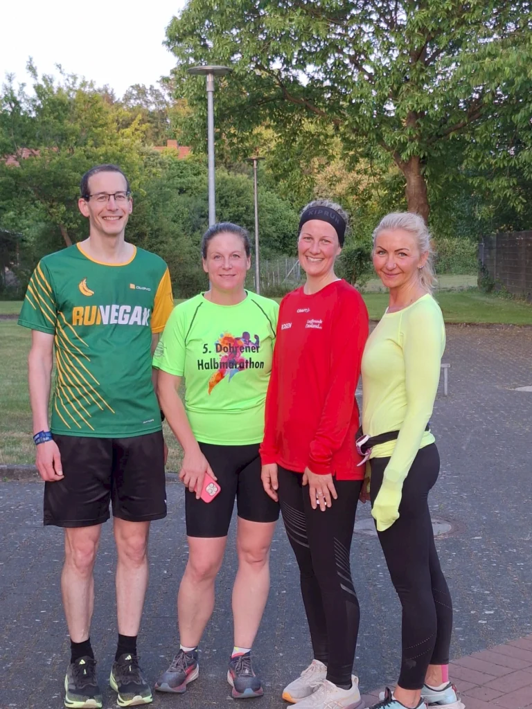 Vier Läufer stehen in Sportkleidung in einem Park, bereit für den 5. Doberner Halbmarathon. Die Gruppe besteht aus zwei Männern und zwei Frauen, die lächeln und motiviert wirken. Im Hintergrund sind Bäume und Laternen zu sehen, was eine sportliche Atmosphäre schafft.
