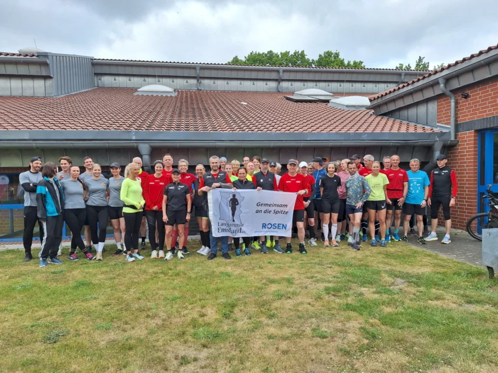 Gruppenfoto von Laufbegeisterten vor einem Vereinsgebäude, die ein Banner mit der Aufschrift „Gemeinsam an die Spitze“ halten. Die Teilnehmer tragen Sportbekleidung in verschiedenen Farben und repräsentieren die Laufgruppe „Lauf Freunde Emsland“.