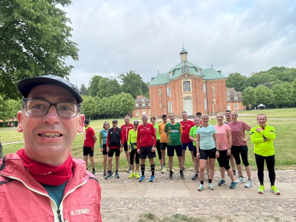 Läufergruppe posiert vor einem historischen Gebäude im Park, ideale Kulisse für ein gemeinsames Lauftraining. Die Teilnehmer tragen sportliche Kleidung und lächeln in die Kamera, während der Fotograf im Vordergrund steht.