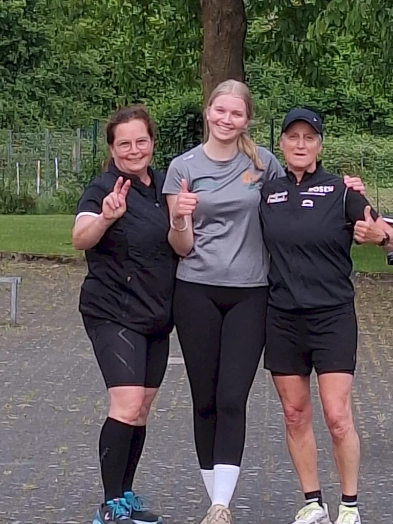 Drei Frauen in Sportbekleidung posieren fröhlich im Freien, zeigen Daumen hoch und ein V-Zeichen. Die Szene spielt sich in einer grünen Umgebung mit Bäumen und Wiese ab, was auf eine sportliche Aktivität oder ein Event hinweist.