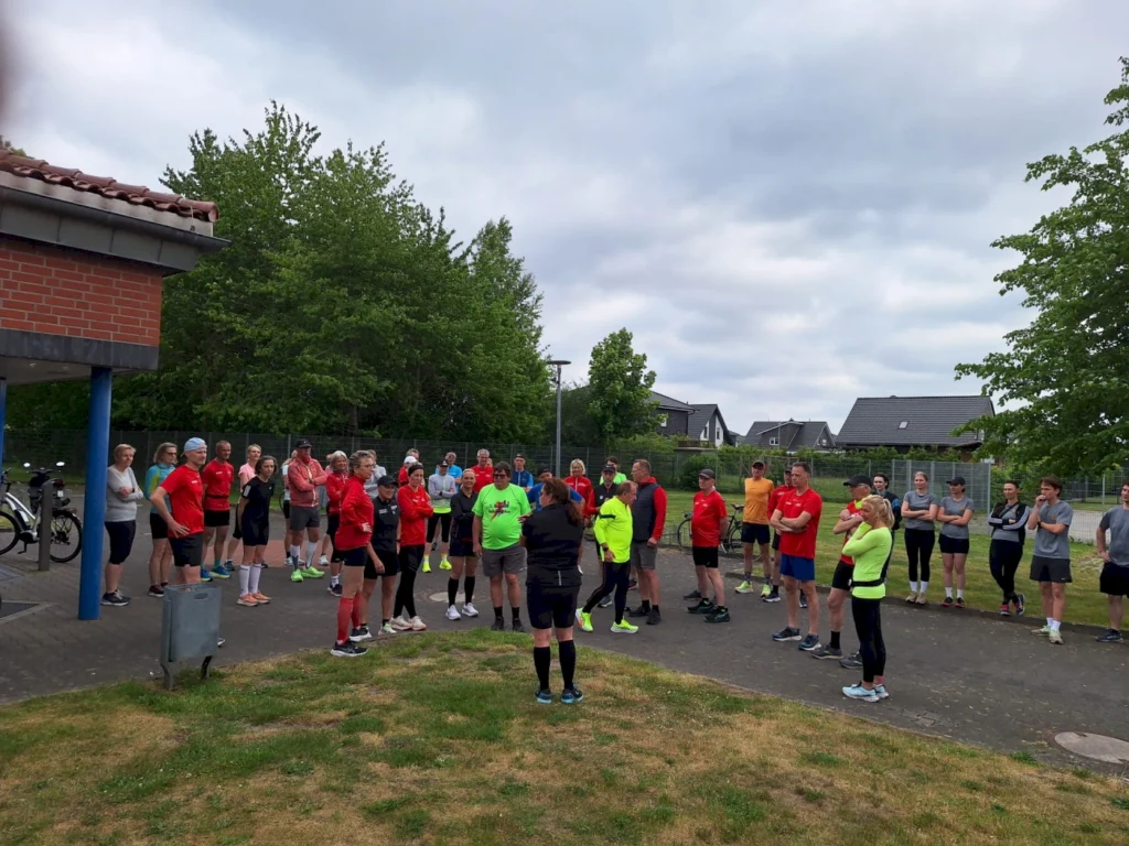 Gruppentraining im Freien mit sportlich gekleideten Teilnehmern, die auf einen Trainer hören. Die Szene zeigt eine Vielzahl von Menschen in Laufkleidung, die sich auf einem Sportgelände versammeln, um an einer Fitnessaktivität teilzunehmen. Im Hintergrund sind Bäume und Wohnhäuser sichtbar.
