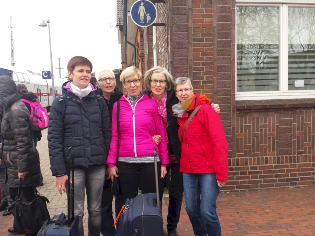 Gruppe von fünf Frauen am Bahnhof, die mit Koffern posieren. Sie tragen warme Kleidung in verschiedenen Farben, darunter eine pinke Jacke. Im Hintergrund sind Züge und ein Fußgängerzeichen sichtbar.