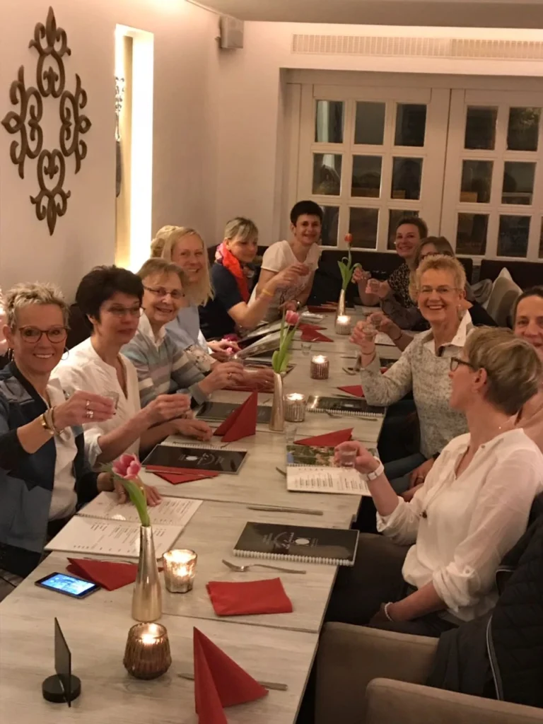 Eine Gruppe von Frauen sitzt an einem festlich gedeckten Tisch in einem Restaurant, hebt Gläser und prostet. Die Tische sind mit roten Servietten und Kerzen dekoriert, während Menüs auf der Tischoberfläche liegen. Die Atmosphäre ist freundlich und gesellig.