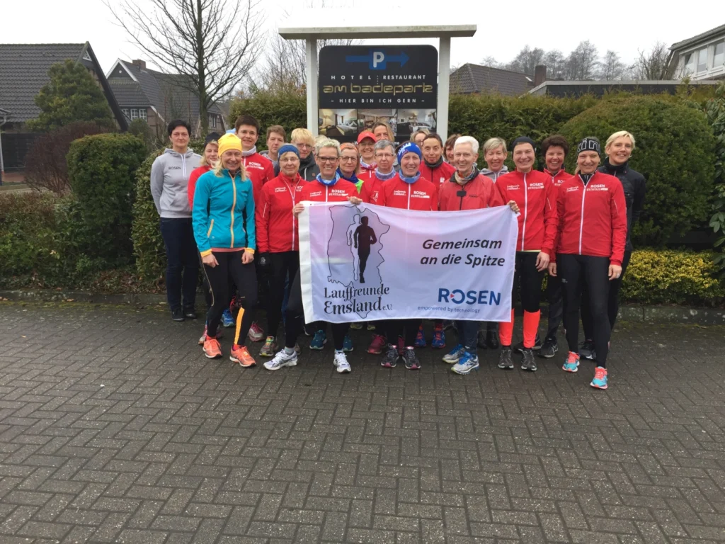 Gruppenfoto von Laufbegeisterten vor dem Hotel am Badepark, die Banner mit der Aufschrift "Lauffreunde Enslade e.V. - Gemeinsam an die Spitze" halten. Die Teilnehmer tragen Sportbekleidung in Rot und anderen Farben und zeigen ihre Begeisterung für das Laufen.