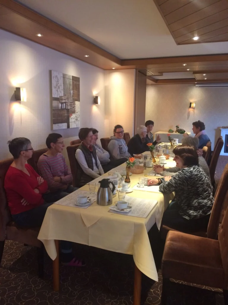 Gruppe von Frauen sitzt an einem Tisch in einem Restaurant, umgeben von gemütlicher Atmosphäre und einem gedeckten Tisch mit Kaffeetassen und Gläsern. Die Wände sind geschmackvoll dekoriert, und es gibt eine kleine Pflanze auf dem Tisch.