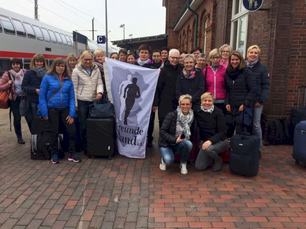 Gruppe von Reisenden am Bahnhof mit Koffern und einer Fahne des Vereins "Freunde der Insel e.V." im Hintergrund, bereit für eine gemeinsame Reise.